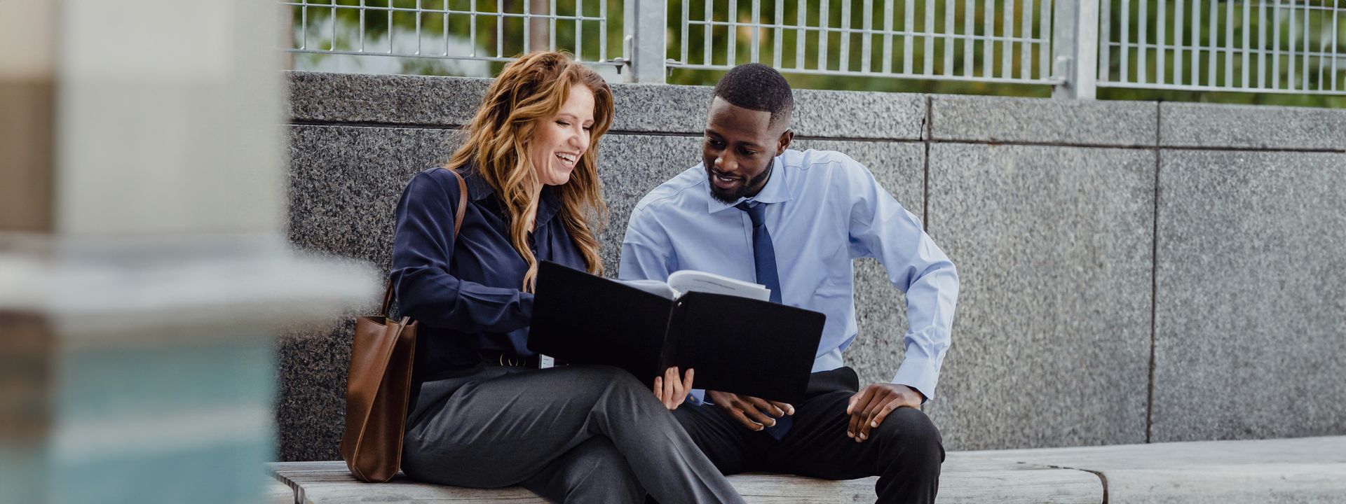 A woman and a man in business attire sit together on an outdoor stone bench, smiling as they review documents in a black folder.