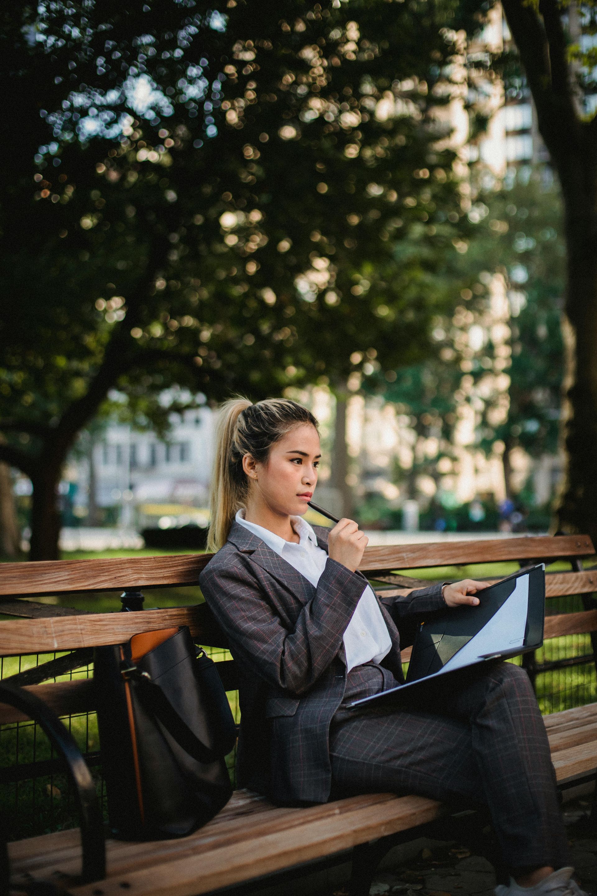 Woman in a grey plaid suit sits thoughtfully on a park bench, holding a pen and a portfolio with documents.