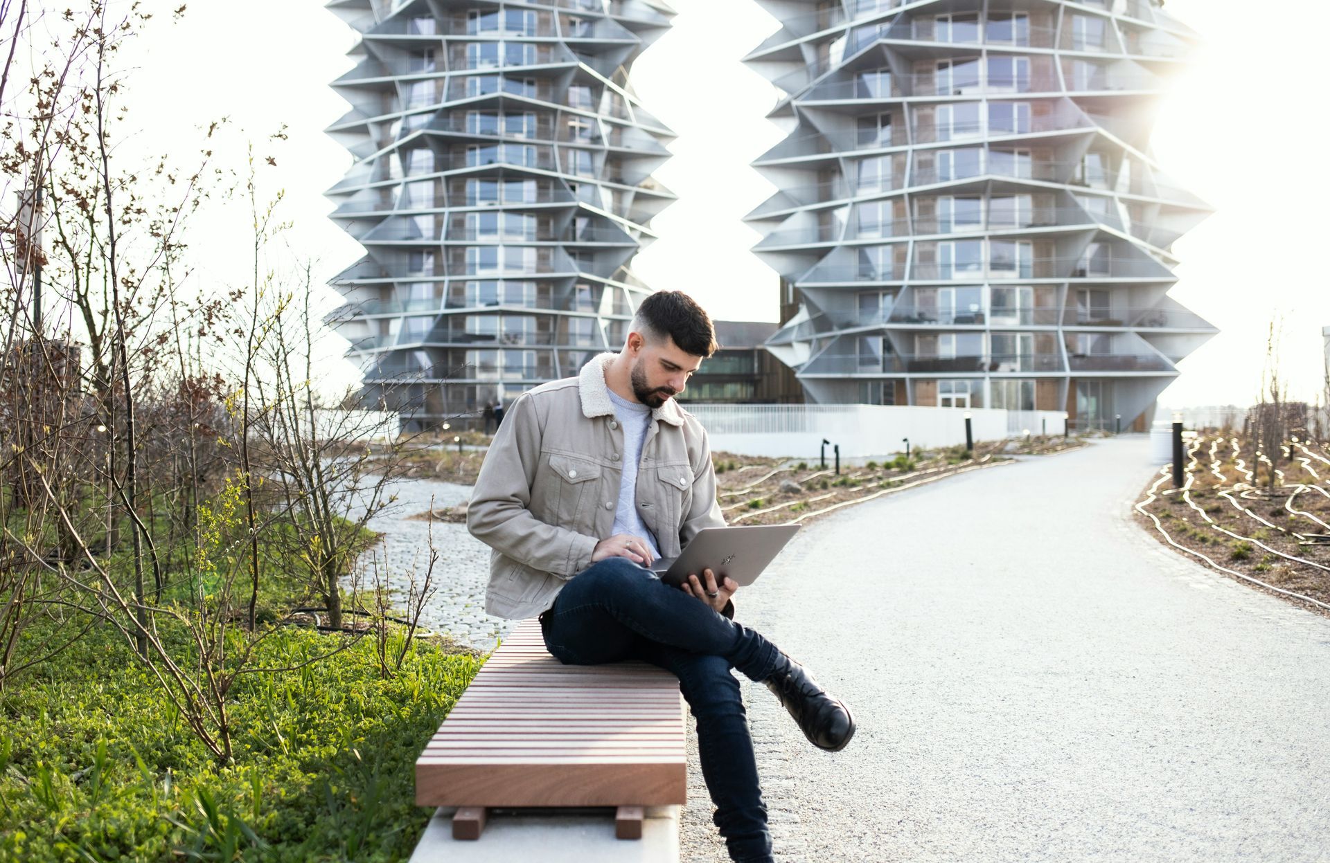 Man sitting on a modern wooden bench outdoors and working on a laptop.