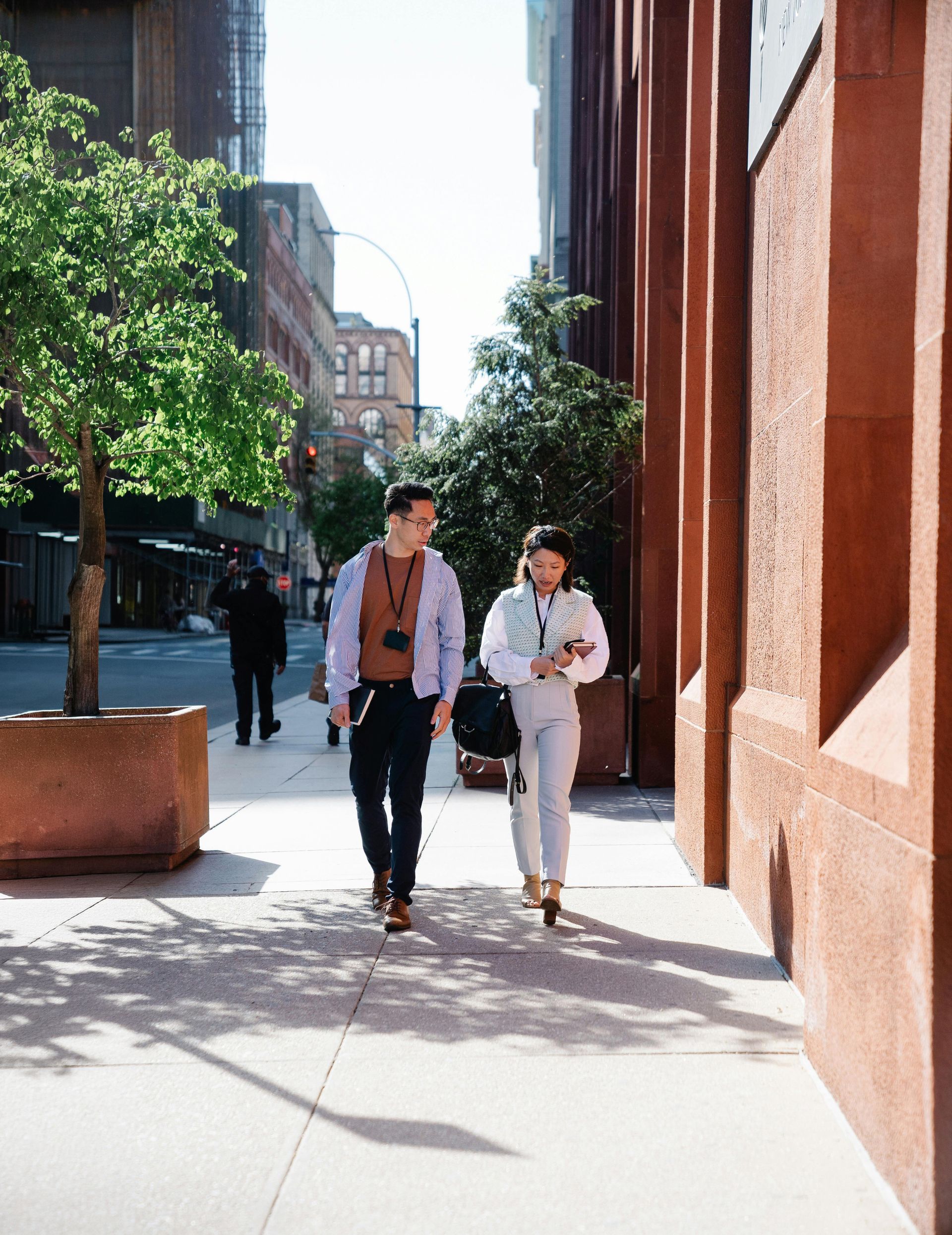Two colleagues walking and talking on a sunny city sidewalk next to a large stone building.