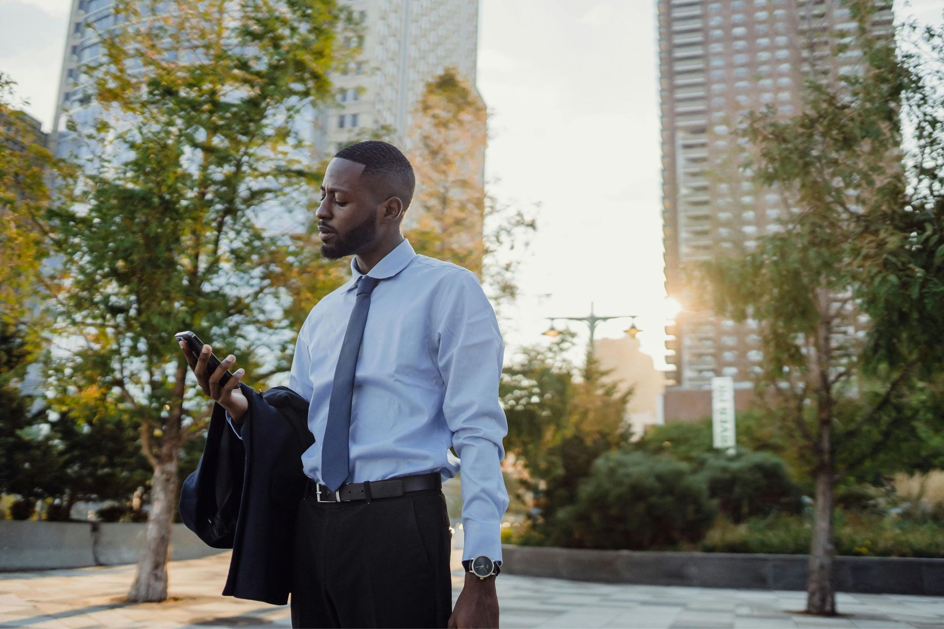 A man in a blue dress shirt and tie looking at his smartphone while standing outdoors with city buildings in the background.