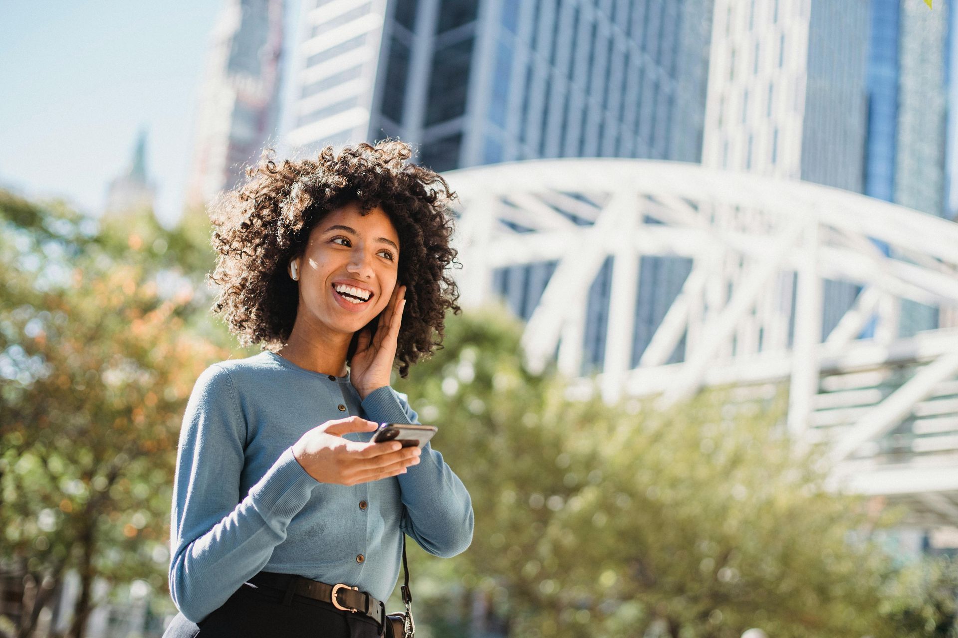 A smiling woman with curly hair holding a smartphone and wearing wireless earbuds in a bright city plaza.