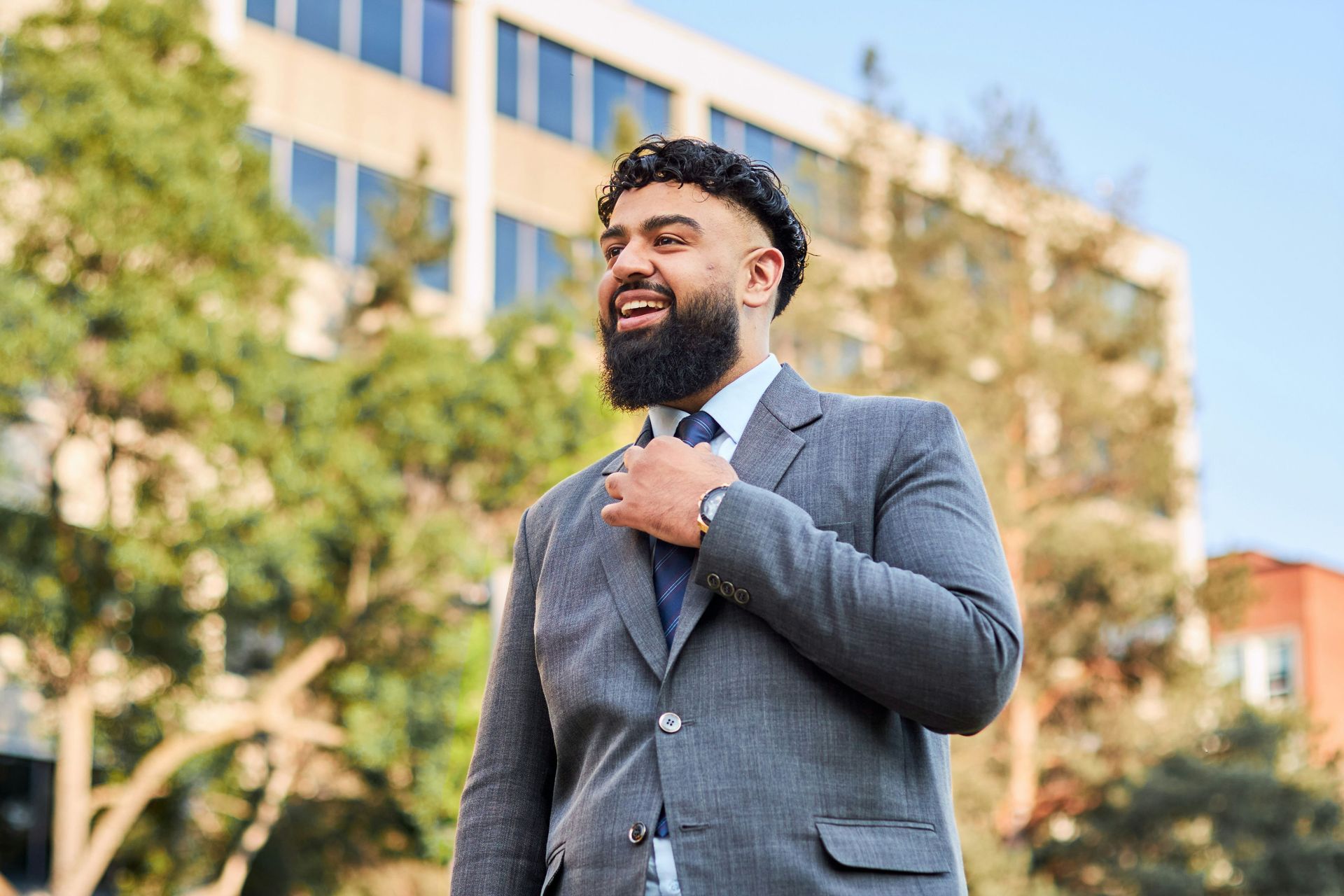 A man in a grey suit and tie smiling and adjusting his collar while standing outside against a backdrop of trees and a building.